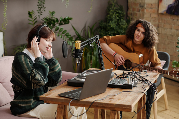A girl in a sweater and headphones sings sitting at a table with equipment on it and a guy with curly hair is sitting next to her and playing guitar